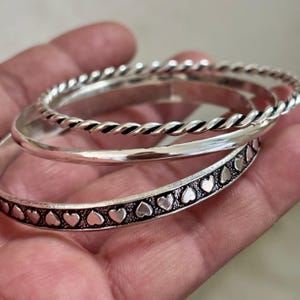 May include: Three silver bangles. One has a twisted rope design, another is plain, and the third features a row of heart-shaped cutouts. The bangles are stacked together, showcasing their different textures and styles.