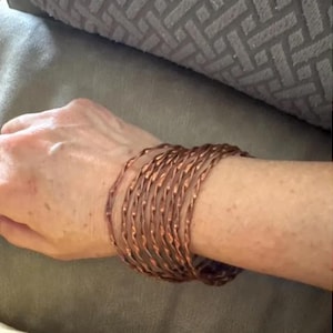 May include: A collection of copper-colored bangles stacked on a wrist. The bangles have a twisted design, creating a textured appearance. The bracelets are a warm, metallic color, reflecting light. The overall style is bohemian and casual.