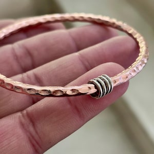 Può includere: Un braccialetto a cerchio color rame con una texture martellata. Il braccialetto presenta una serie di anelli color argento come elemento decorativo. Il braccialetto è un gioiello.