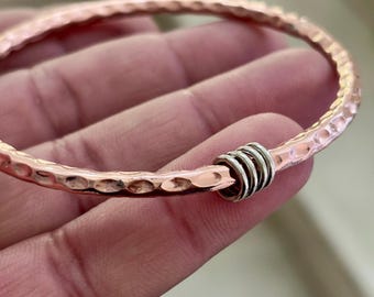 May include: A copper-coloured bangle bracelet with a hammered texture. The bracelet has a series of silver-coloured rings as a decorative element. The bracelet is a piece of jewellery.