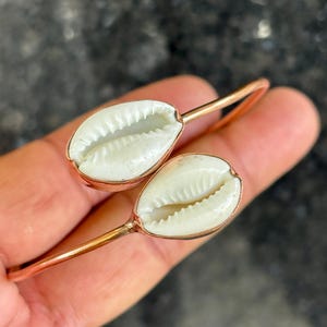 Könnte beinhalten: Ein kupferfarbenes Armband in offener Form mit zwei weißen Kaurimuscheln an den Enden. Die ovalen Muscheln weisen einen zentralen Schlitz und gezackte Ränder auf und sind in einem glatten, metallischen Rahmen eingefasst, der den Armreif bildet.
