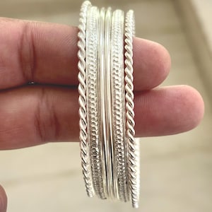 May include: A collection of silver bangles with various textures, including twisted rope and dotted patterns. The bangles are stacked together, showcasing their different designs and shiny finish.
