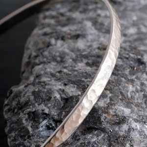 Op de afbeelding: Een zilveren gehammerde armband. De armband is gemaakt van glad, gepolijst metaal en heeft een gehammerde textuur. De armband wordt getoond op een grijs, getextureerd oppervlak.