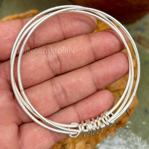 May include: Three silver bangles, held in a hand, are the focus of this image. The bangles are thin and round, with a decorative wire wrap detail. The jewelry is displayed against a natural, textured background.