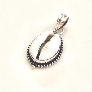 May include: A silver pendant with a white cowrie shell. The shell is set in a detailed silver frame with a beaded border. The pendant has a loop for a chain.