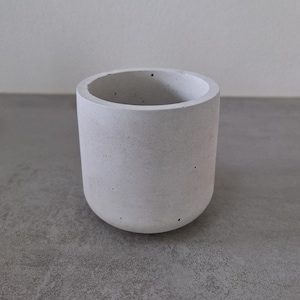 Pode incluir: Um pequeno vaso de cimento branco com uma superfície áspera e texturizada. O vaso é redondo e tem uma borda ligeiramente alargada.