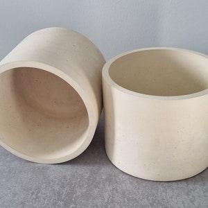 May include: Two off-white concrete planters, one slightly larger than the other, with a rough, textured surface. The planters are empty and ready for plants.