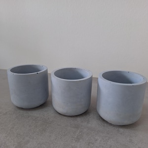 Può includere: Tre piccoli vasi in cemento blu chiaro. I vasi sono rotondi e hanno una superficie leggermente ruvida.