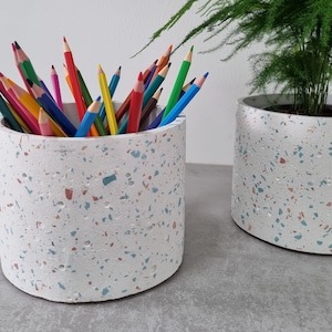 Peut inclure: Deux pots en terrazzo blanc, l'un avec un bouquet de crayons de couleur et l'autre avec une plante verte.