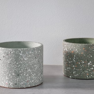 Könnte beinhalten: Zwei runde Terrazzo-Blumentöpfe in hellgrün mit weißen und orangefarbenen Flecken. Die Töpfe stehen auf einer grauen Oberfläche.