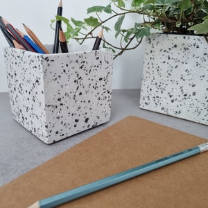 May include: A white ceramic pencil holder with black flecks, filled with pencils. The holder is sitting on a gray surface next to a brown notebook and a blue pencil.