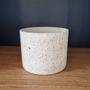 Può includere: Un vaso cilindrico bianco con una finitura in terrazzo macchiettato. Il vaso è in cemento e ha un bordo liscio e arrotondato.