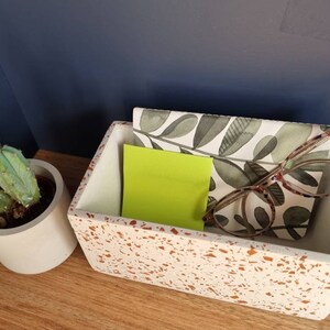 May include: A white rectangular terrazzo tray with a green and white patterned paper inside. The tray contains a yellow sticky note and a pair of spectacles.
