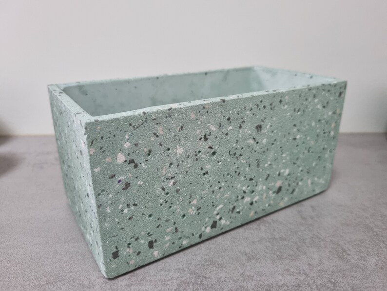 May include: A rectangular terrazzo planter in a light green colour with white and grey flecks. The planter is made of concrete and has a smooth finish.