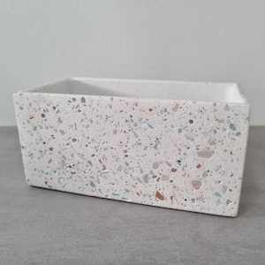 Könnte beinhalten: Ein rechteckiger weißer Pflanzer mit einem Terrazzo-Muster. Der Pflanzer hat ein gesprenkeltes Design mit kleinen blauen, rosa und orangen Spänen.