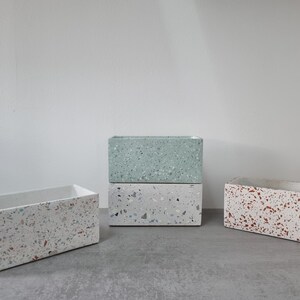 以下が含まれることがあります： 異なる色の長方形のテラゾプランター3つ。プランターは白を基調とし、青、緑、赤の斑点が入っています。