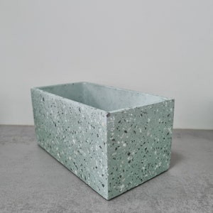 May include: A light green rectangular planter with a speckled terrazzo finish. The planter is made of concrete and has a smooth, slightly textured surface.