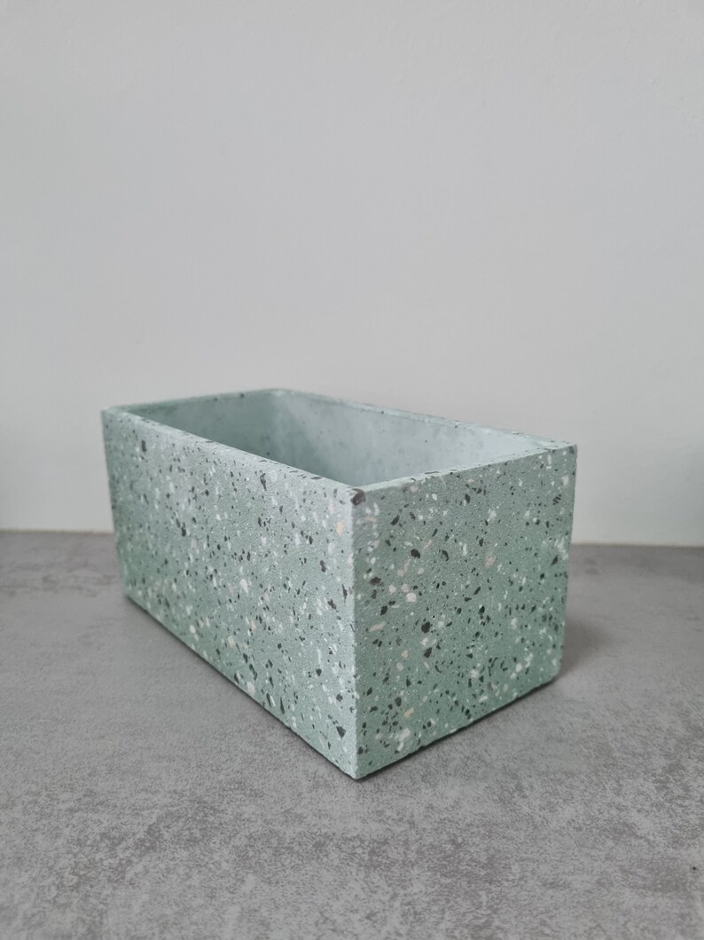 May include: A light green rectangular planter with a speckled terrazzo finish. The planter is made of concrete and has a smooth, slightly textured surface.