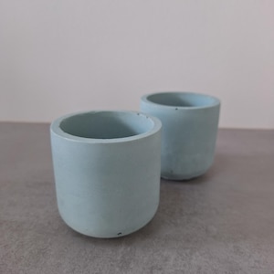 Op de afbeelding: Twee kleine, lichtblauwe betonnen plantenpotten. De cilindrische potten hebben een afgeronde basis en een gladde, matte afwerking. De plantenbakken zijn leeg en klaar voor kleine planten of vetplanten. De achtergrond is neutraal grijs.