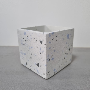 Op de afbeelding: Een witte vierkante plantenbak met een terrazzo-patroon in zwart en lichtblauw. De plantenbak is gemaakt van beton en heeft een ruwe textuur.