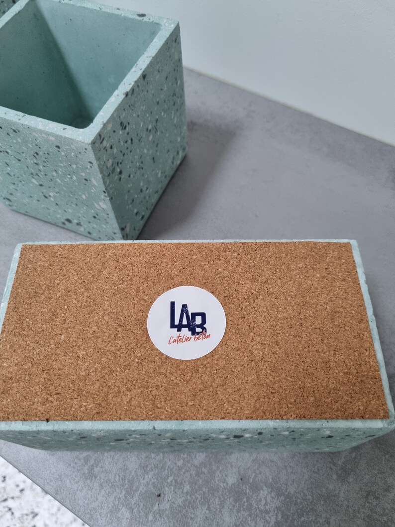 May include: A light blue concrete planter with a speckled finish. The planter has a cork base and a white sticker with the text "LAB L'atelier Beton".