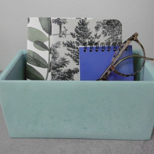 May include: A light blue concrete rectangular tray with three notebooks and a pair of glasses inside. The notebooks have a black and white floral pattern and a blue cover. The glasses are tortoiseshell.