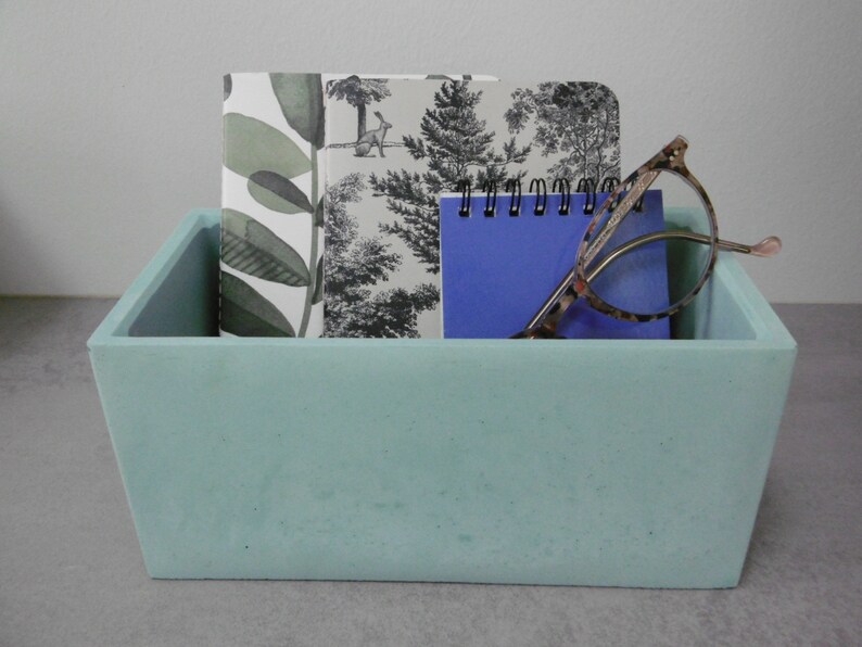 May include: A light blue concrete rectangular tray with three notebooks and a pair of glasses inside. The notebooks have a black and white floral pattern and a blue cover. The glasses are tortoiseshell.