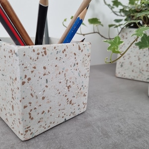 May include: A square, speckled pencil holder made of light-colored concrete. The holder contains several pencils in various colors. A matching planter and candle holder are in the background, with green ivy.