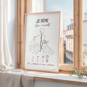 Un bonhomme bâton minimaliste et esthétique sème des graines dans un jardin. Il disperse soigneusement des graines. Des fleurs épanouies et d’autres en train d’éclore illustrent le titre ‘Je sème la réussite’. Affiche encadrée en bois posée sur un rebord de fenêtre orné de rideaux en lin beige. Le cadre est légèrement incliné.