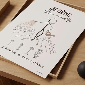 Un bonhomme bâton minimaliste et esthétique sème des graines dans un jardin. Il disperse soigneusement des graines. Des fleurs épanouies et d’autres en train d’éclore illustrent le titre ‘Je sème la réussite’. Affiche agrandie posée sur un cadre en bois, lui-même posé sur une table.