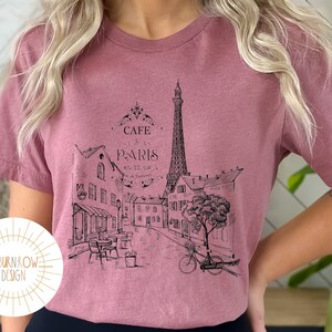 May include: A mauve t-shirt with a black and white graphic print of the Eiffel Tower and a Parisian street scene. The graphic includes the text "Cafe Paris".