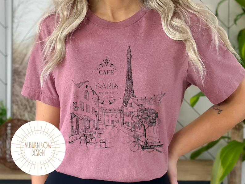 May include: A mauve t-shirt with a black and white graphic print of the Eiffel Tower and a Parisian street scene. The graphic includes the text "Cafe Paris".
