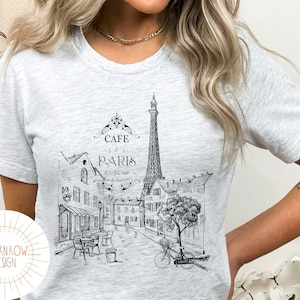 May include: A heather grey t-shirt with a black and white illustration of the Eiffel Tower and a Parisian street scene. The text "Cafe de Paris" is printed on the shirt.