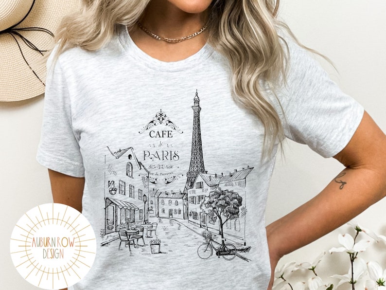 May include: A heather grey t-shirt with a black and white illustration of the Eiffel Tower and a Parisian street scene. The text "Cafe de Paris" is printed on the shirt.