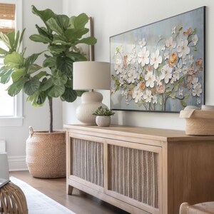 Pu&ograve; includere: Un mobile in legno chiaro con due ante che presentano un design intrecciato. Il mobile &egrave; decorato con una lampada in ceramica bianca con un paralume color crema e un dipinto floreale sopra. Una grande pianta in vaso con foglie verdi si trova a sinistra del mobile.