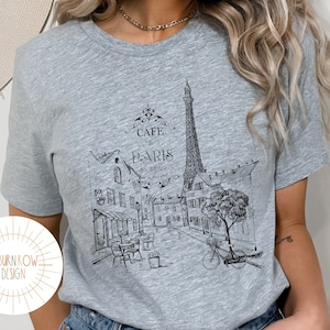 May include: A gray t-shirt with a black and white illustration of the Eiffel Tower and a Parisian street scene. The text "Cafe de Paris" is printed on the shirt.