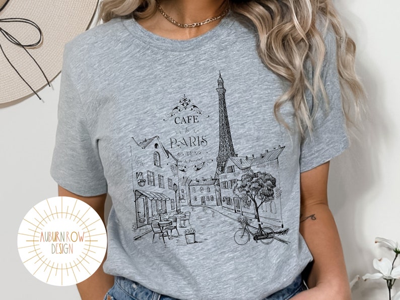 May include: A gray t-shirt with a black and white illustration of the Eiffel Tower and a Parisian street scene. The text "Cafe de Paris" is printed on the shirt.