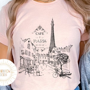 May include: A pink t-shirt with a black and white illustration of the Eiffel Tower and Parisian buildings. The text "Cafe de Paris" is printed on the shirt.