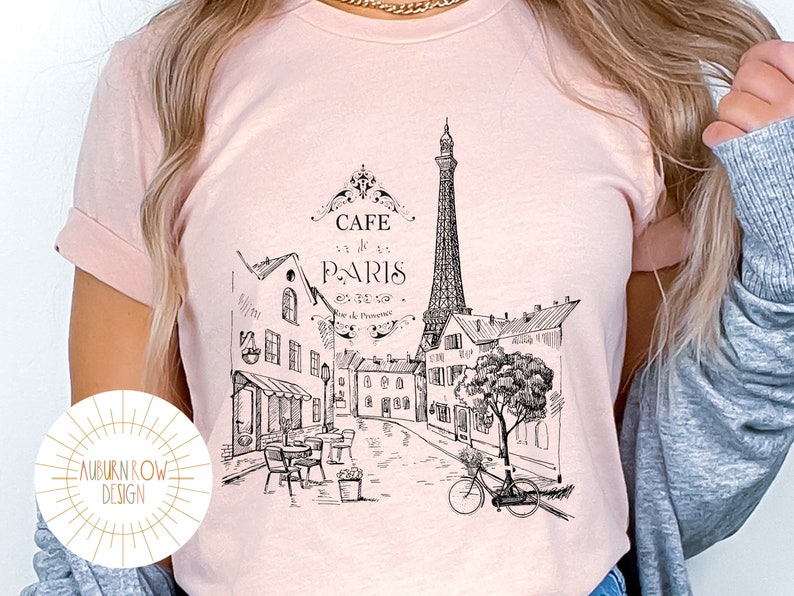 May include: A pink t-shirt with a black and white illustration of the Eiffel Tower and Parisian buildings. The text "Cafe de Paris" is printed on the shirt.