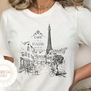May include: White t-shirt with a black and white illustration of a Parisian street scene. The illustration includes the Eiffel Tower, buildings, a cafe, a bicycle, and a tree. The text "Cafe de Paris" is written above the illustration.