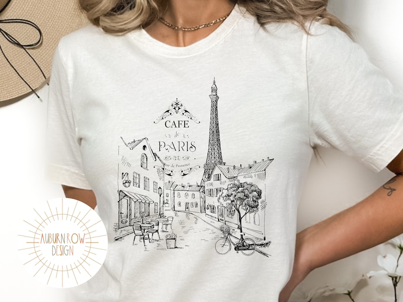 May include: White t-shirt with a black and white illustration of a Parisian street scene. The illustration includes the Eiffel Tower, buildings, a cafe, a bicycle, and a tree. The text "Cafe de Paris" is written above the illustration.