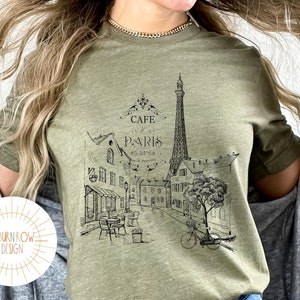 May include: A green t-shirt with a black and white graphic of the Eiffel Tower and a Parisian street scene. The text "Cafe de Paris" is printed on the shirt.