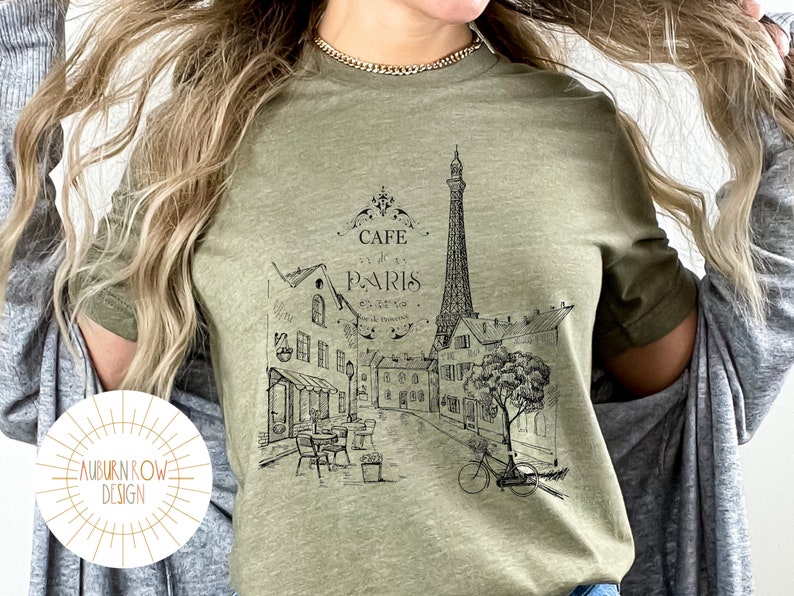 May include: A green t-shirt with a black and white graphic of the Eiffel Tower and a Parisian street scene. The text "Cafe de Paris" is printed on the shirt.
