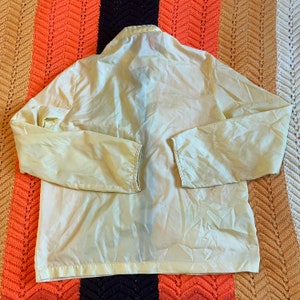 May include: A pale yellow lightweight jacket with a zipper closure and a high collar. The jacket is made of a thin, smooth fabric and has a relaxed fit.