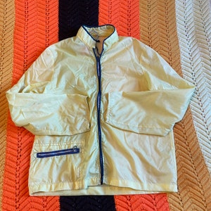 May include: A light yellow windbreaker jacket with a zipper closure and a blue zipper pull. The jacket has a stand-up collar and a single pocket on the left side.