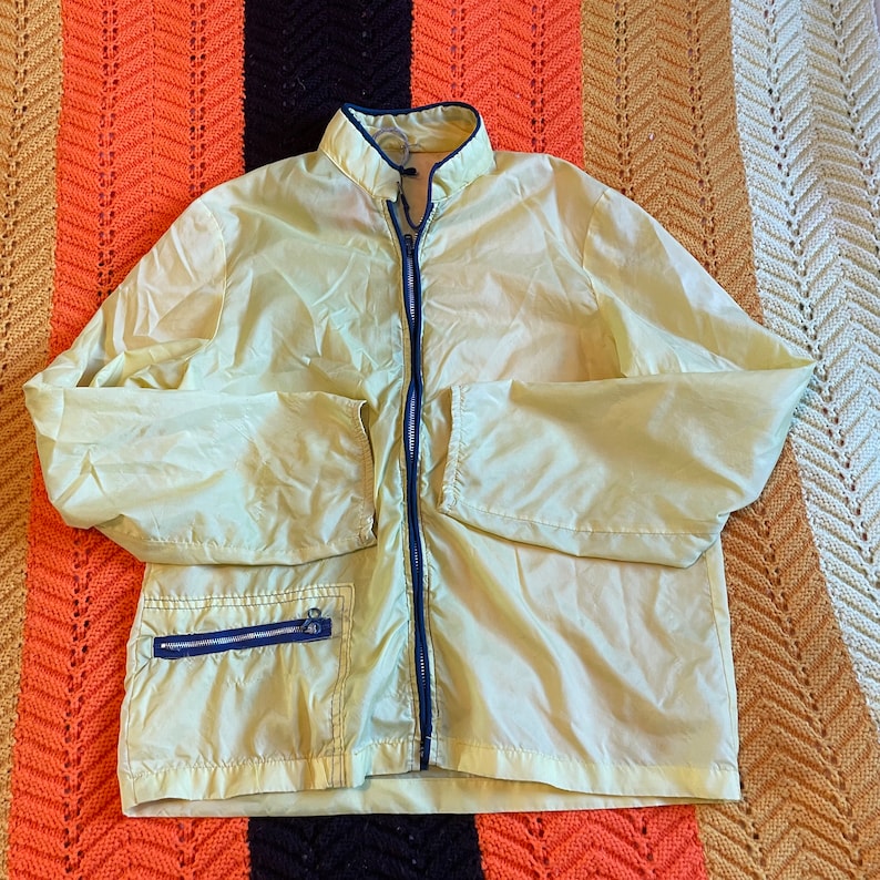 May include: A light yellow windbreaker jacket with a zipper closure and a blue zipper pull. The jacket has a stand-up collar and a single pocket on the left side.
