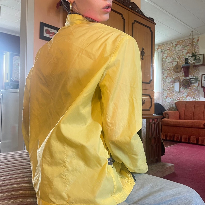 May include: A yellow, lightweight jacket with a zipper closure. The jacket is made of a smooth, silky fabric and has a relaxed fit.