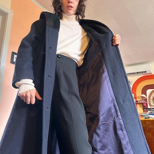 May include: A long, navy blue coat with a dark green collar and a purple interior lining. The coat is buttoned up and appears to be made of a heavy fabric. The coat is being held open, revealing the lining.