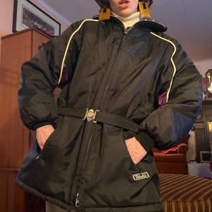 May include: Black ski jacket with white and purple trim, a belt with a silver buckle, and the word "Nikoli" in white lettering on the front.