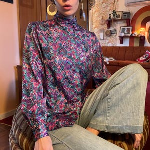Puede incluir: Una mujer lleva una blusa de manga larga y cuello alto con estampado floral de colores. La blusa está hecha de una tela sedosa y tiene un corte holgado. La mujer también lleva pantalones vaqueros azules.